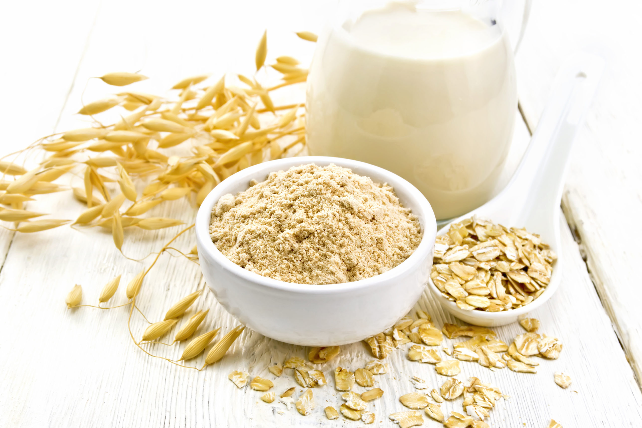 Flour oat in bowl, milk in a jug, oatmeal in spoon, oaten stalks on the background of light wooden board